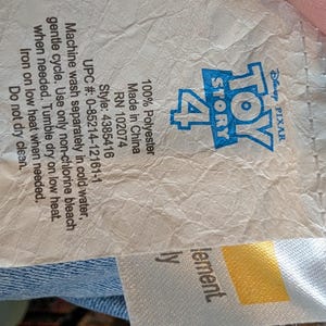 May include: A white fabric care label for a Toy Story 4 product. The label features the blue Toy Story 4 logo, 100% Polyester material content, style number 4385416, UPC 0-85214-12161-1, and care instructions including machine wash cold and tumble dry low.