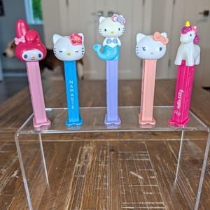 May include: Five Pez candy dispensers featuring character heads including a pink My Melody, a blue Hello Kitty with Namaste text, a mermaid Hello Kitty on a purple stem, a white Hello Kitty with a peach bow, and a white unicorn on a pink stem.