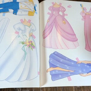 May include: A children's paper doll book open to a page with illustrations of dresses and a prince's outfit. The dresses are in shades of pink and white, with a blue cape. The prince's outfit is blue and gold.