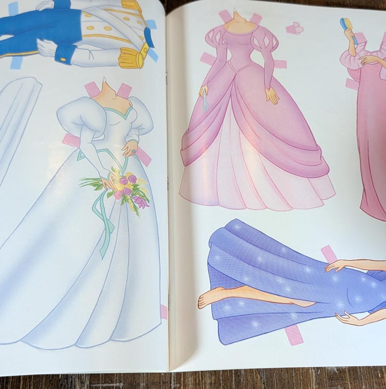 May include: A children's paper doll book open to a page with illustrations of dresses and a prince's outfit. The dresses are in shades of pink and white, with a blue cape. The prince's outfit is blue and gold.
