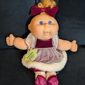 May include: A Cabbage Patch Kids doll, Terry Claire, with blonde hair and blue eyes. She wears a burgundy velvet dress with a white fur trim and a matching bow. The doll has a tag that reads "Terry Claire, My Birthday is December 5, I like toy boats!"