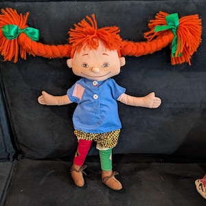 Op de afbeelding: Een pluchen pop met feloranje garen vlechten, vastgebonden met groene strikken. De pop draagt een blauw shirt, een korte broek met patroon en mismatched rode en groene leggings. Bruine schoenen maken de outfit compleet. Het gezicht van de pop heeft sproeten en een zachte glimlach.