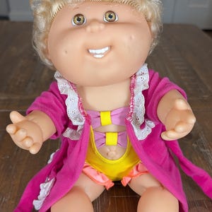 May include: A doll with blonde curly hair and brown eyes wearing a bright pink robe with white lace trim over a yellow leotard featuring two pink and yellow bows on the front. The doll has a smiling expression with visible teeth and dimples.