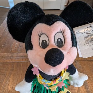May include: A Minnie Mouse plush doll with black ears, a pink face, and a yellow and green hula skirt. The doll has a tag that reads "Made Exclusively For The Disney Store". The doll is wearing a lei with pink flower accents.