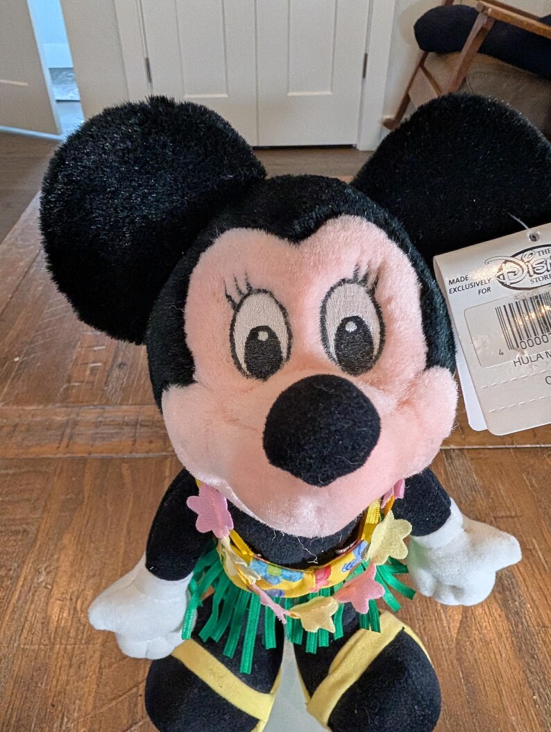 May include: A Minnie Mouse plush doll with black ears, a pink face, and a yellow and green hula skirt. The doll has a tag that reads "Made Exclusively For The Disney Store". The doll is wearing a lei with pink flower accents.