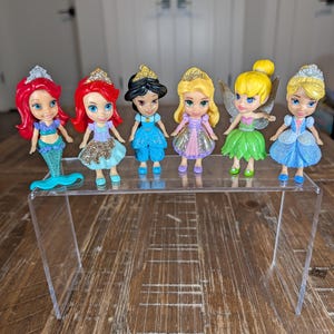 May include: A set of six small plastic character figurines featuring various princesses and a fairy. The figures have glittery accents on their outfits and crowns, with distinct hair colors including red, black, blonde, and yellow, standing on a clear stand.