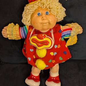 May include: A vintage clown doll with a yellow hat and curly blonde hair. The doll wears a red outfit with colorful heart accents, a clown collar, and red shoes. The doll's arms have rainbow sleeves and gold cuffs.
