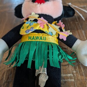 May include: A plush toy of a cartoon character wearing a yellow and green Hawaiian outfit. The outfit includes a yellow top with blue floral designs, a green grass skirt, and a headband with pink and yellow flowers. The word "HAWAII" is printed on the outfit.