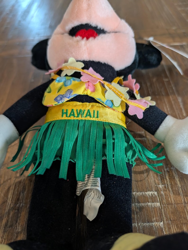May include: A plush toy of a cartoon character wearing a yellow and green Hawaiian outfit. The outfit includes a yellow top with blue floral designs, a green grass skirt, and a headband with pink and yellow flowers. The word "HAWAII" is printed on the outfit.