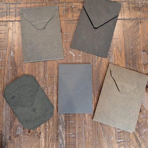 May include: Five textured, envelope-style paper pouches in shades of green and tan are arranged on a wooden surface. Each pouch has a flap closure. The pouches vary slightly in shape and color, creating a natural, earthy aesthetic.