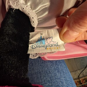 Peut inclure: Une peluche avec un bras noir et une bordure en dentelle blanche. Le jouet porte une &eacute;tiquette indiquant "Disneyland Resort" avec un ch&acirc;teau et une illustration de cochon rose. Le jouet est sur une surface en jean.