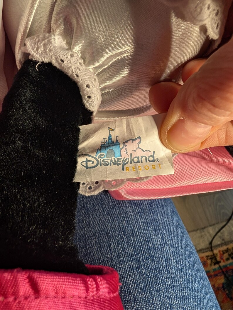 Peut inclure: Une peluche avec un bras noir et une bordure en dentelle blanche. Le jouet porte une &eacute;tiquette indiquant "Disneyland Resort" avec un ch&acirc;teau et une illustration de cochon rose. Le jouet est sur une surface en jean.
