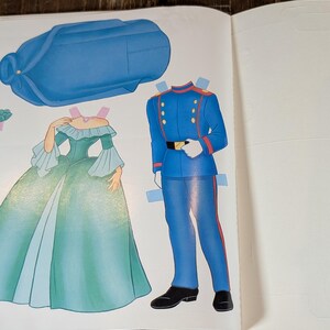 May include: A vintage paper doll page featuring a blue hat, a green and blue gown, and a blue military uniform. The gown has a ruffled neckline and bell sleeves. The uniform has gold buttons and a black belt. The paper dolls are cut out.