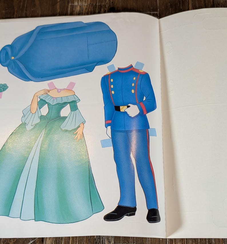 May include: A vintage paper doll page featuring a blue hat, a green and blue gown, and a blue military uniform. The gown has a ruffled neckline and bell sleeves. The uniform has gold buttons and a black belt. The paper dolls are cut out.