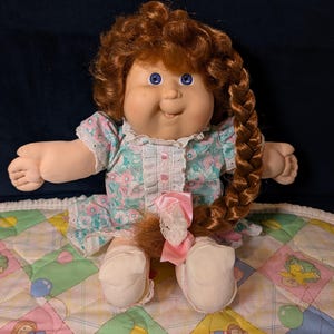 May include: A vintage doll with red hair styled in a braid and a floral dress. The doll has blue eyes and is seated on a colorful quilt with cartoon characters. A pink bow adorns the end of the braid.
