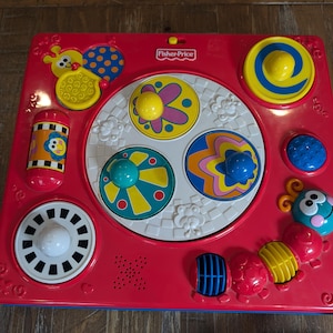 May include: A red Fisher-Price activity center with various colorful interactive elements. Features include spinning wheels, push buttons, and a variety of shapes and textures. The toy is designed for early childhood development and play.