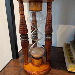 May include: A wooden hourglass with three glass bulbs filled with sand. The hourglass is brown with a reddish-brown stain on the base.