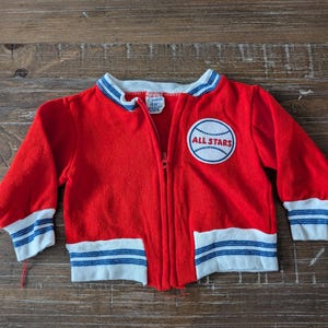 May include: Red zippered jacket with blue and white stripes on the cuffs and waistband. The jacket has a patch on the front that says "ALL STARS" with a baseball graphic.