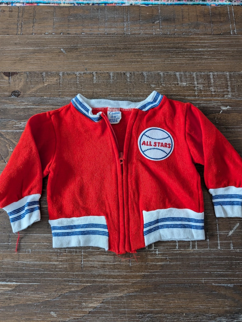 May include: Red zippered jacket with blue and white stripes on the cuffs and waistband. The jacket has a patch on the front that says "ALL STARS" with a baseball graphic.