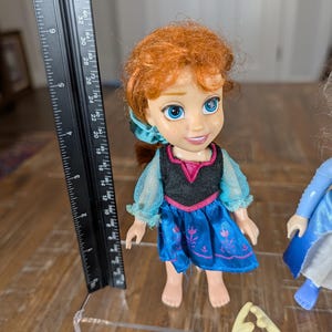 May include: A doll of a character with red hair, wearing a blue dress with a black bodice and a pink neckline. The doll is approximately 5 inches tall. A ruler is next to the doll.