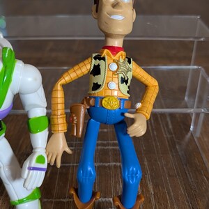 May include: A plastic action figure of a cowboy character with brown hair, wearing a yellow long-sleeve shirt with a black cow-print vest, a gold sheriff badge, a blue belt with a gold buckle, blue pants, brown boots, and a brown holster.
