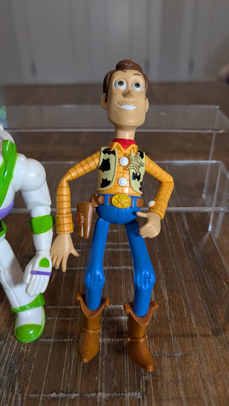 May include: A plastic action figure of a cowboy character with brown hair, wearing a yellow long-sleeve shirt with a black cow-print vest, a gold sheriff badge, a blue belt with a gold buckle, blue pants, brown boots, and a brown holster.