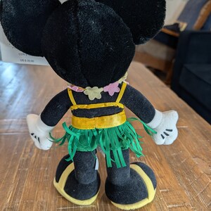 May include: A black plush Minnie Mouse doll wearing a yellow and green hula skirt, a yellow top, and a pink and yellow flower necklace. The doll has white gloves and yellow accents on the shoes. The doll's ears are large and round.