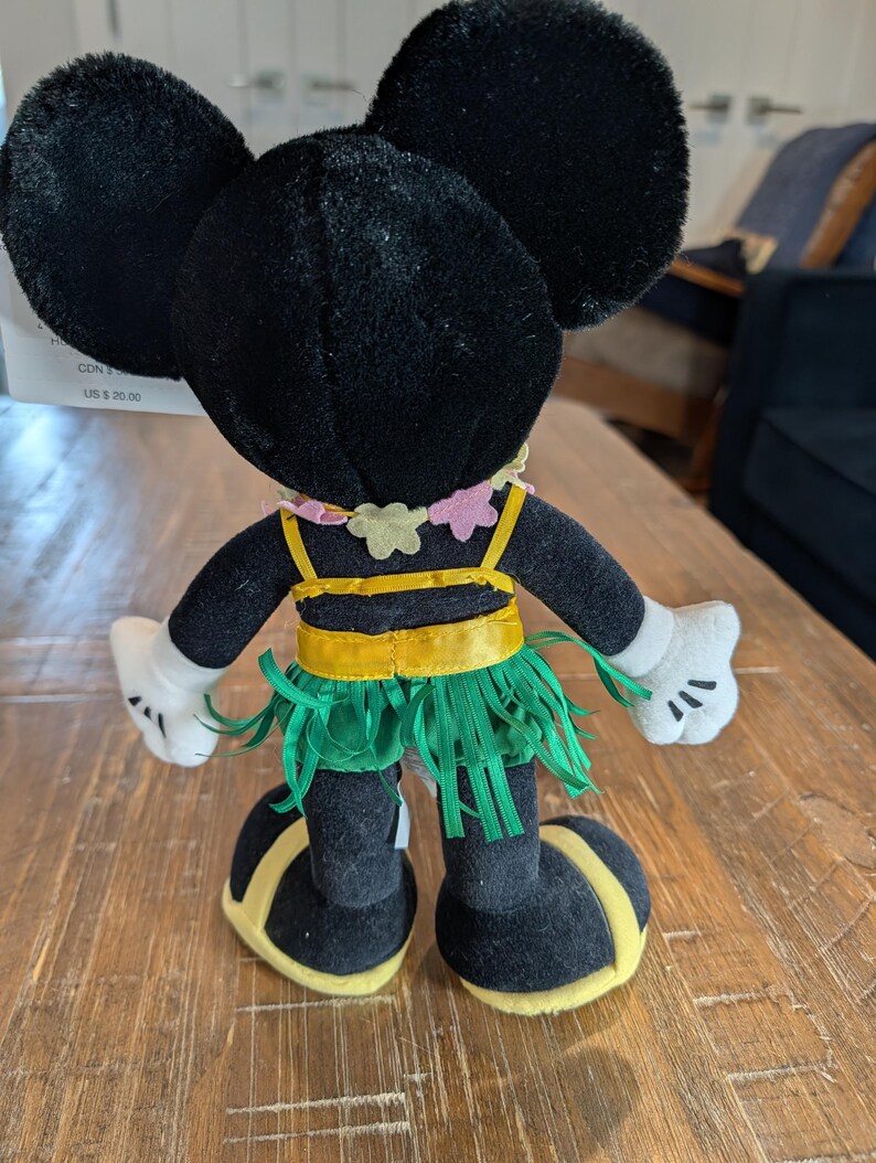 May include: A black plush Minnie Mouse doll wearing a yellow and green hula skirt, a yellow top, and a pink and yellow flower necklace. The doll has white gloves and yellow accents on the shoes. The doll's ears are large and round.
