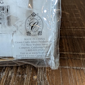 May include: A close-up of a product package with text. The package is sealed in clear plastic. The text includes "MADE IN CHINA", the Crown Crafts logo, and contact information for Crown Crafts Infant Products.
