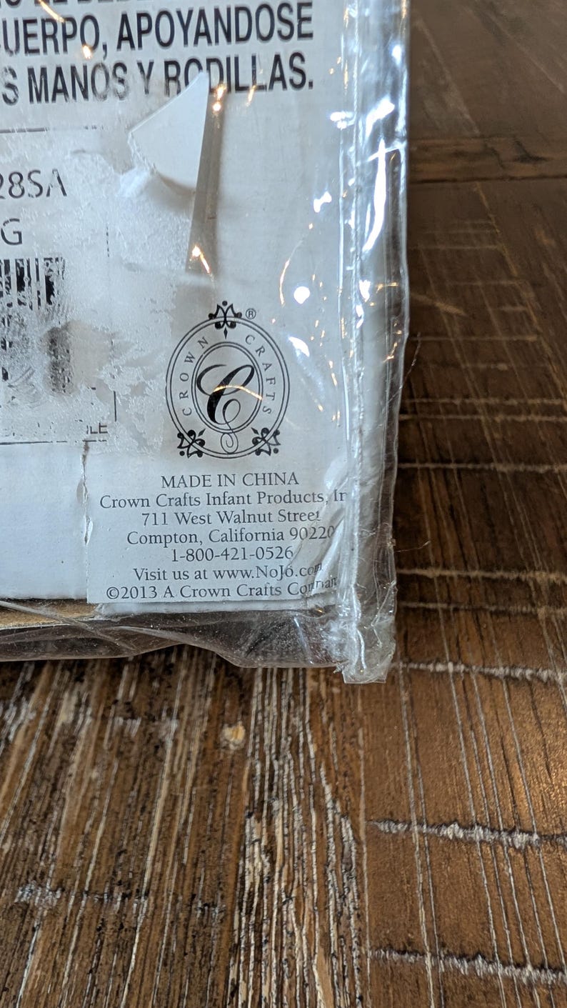 May include: A close-up of a product package with text. The package is sealed in clear plastic. The text includes "MADE IN CHINA", the Crown Crafts logo, and contact information for Crown Crafts Infant Products.