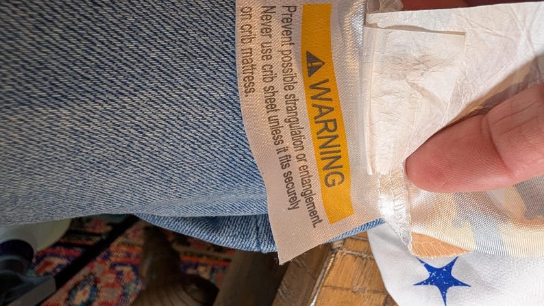 May include: A light blue fabric item featuring a white care label with a yellow warning banner. The text reads: WARNING Prevent possible strangulation or entanglement. Never use crib sheet unless it fits securely. A white fabric with blue stars.