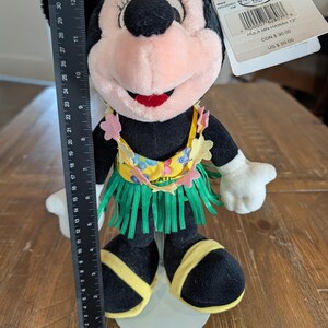 May include: A Minnie Mouse plush doll, approximately 12 inches tall, dressed in a Hawaiian-themed outfit. The doll features a black body, pink face, and a grass skirt. The doll has a yellow top with floral accents and is standing on a white base.
