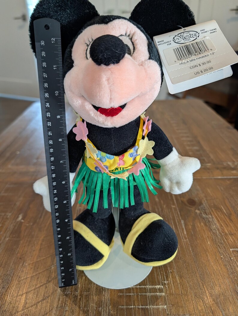 May include: A Minnie Mouse plush doll, approximately 12 inches tall, dressed in a Hawaiian-themed outfit. The doll features a black body, pink face, and a grass skirt. The doll has a yellow top with floral accents and is standing on a white base.