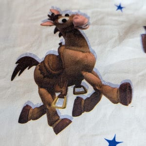 May include: A white fabric featuring a repeating pattern of cartoon characters including a brown horse with a saddle and stirrups, a white spork character with red arms, and small blue stars scattered across the surface of the material.