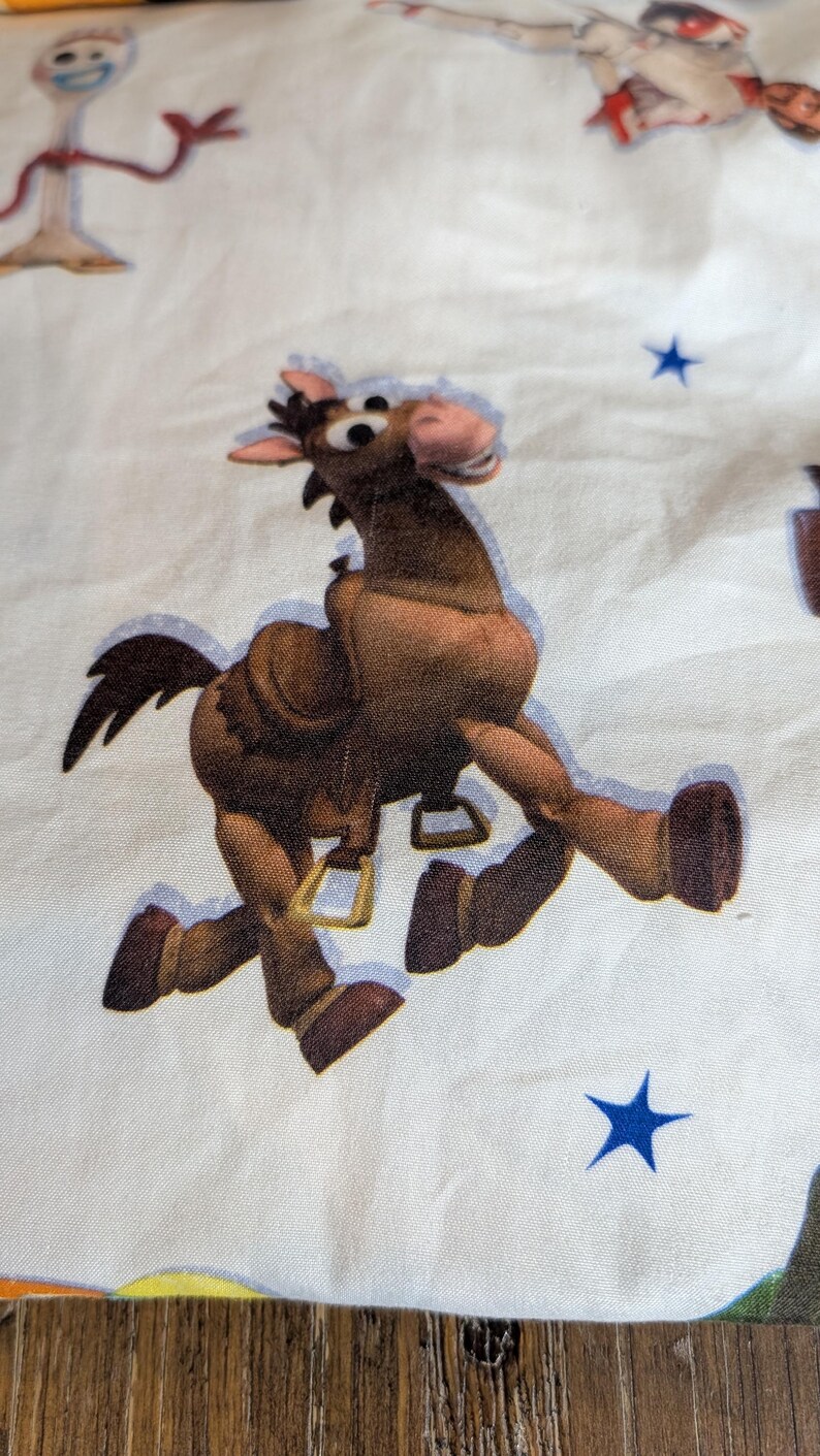 May include: A white fabric featuring a repeating pattern of cartoon characters including a brown horse with a saddle and stirrups, a white spork character with red arms, and small blue stars scattered across the surface of the material.