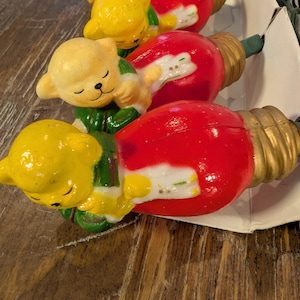 May include: A string of Christmas lights featuring yellow teddy bear figures wearing green and white outfits, paired with large red bulbs. The bulbs have gold-colored bases. The lights are arranged on a white cardboard backing.