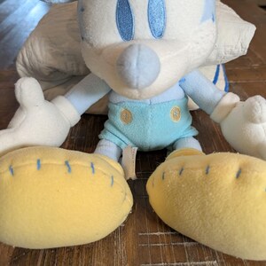 Op de afbeelding: Een pluche Mickey Mouse-speelgoed in lichtblauw, wit en geel. Het speelgoed heeft blauwe stikseldetails, blauwe ogen en gele voeten. Het speelgoed staat op een houten oppervlak.