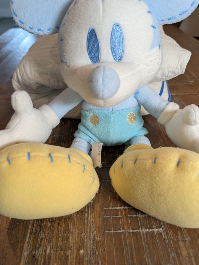 Op de afbeelding: Een pluche Mickey Mouse-speelgoed in lichtblauw, wit en geel. Het speelgoed heeft blauwe stikseldetails, blauwe ogen en gele voeten. Het speelgoed staat op een houten oppervlak.