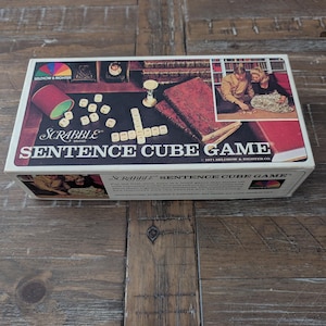 May include: A vintage Scrabble Sentence Cube Game box. The box features a colorful illustration of the game's components, including dice, books, and a couple playing the game. The game's name is prominently displayed in large white letters.