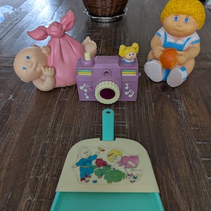 May include: A set of vintage Cabbage Patch Kids toys, including a baby doll in a pink swaddle, a doll holding a ball, and a camera toy. There is also a small plastic dustpan with a picture of two Cabbage Patch Kids on it.