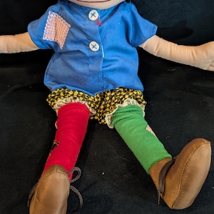 Op de afbeelding: Handgemaakte pop met rood garen haar, sproeten en een lachend gezicht. Ze draagt een blauw shirt, een korte broek met patroon en mismatched rode en groene sokken. Bruine schoenen met veters maken de outfit compleet. Een grillig, handgemaakt item.