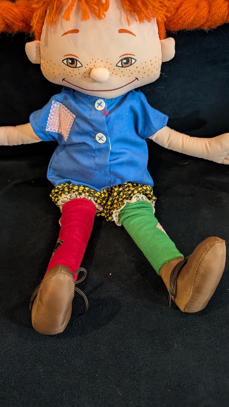 Op de afbeelding: Handgemaakte pop met rood garen haar, sproeten en een lachend gezicht. Ze draagt een blauw shirt, een korte broek met patroon en mismatched rode en groene sokken. Bruine schoenen met veters maken de outfit compleet. Een grillig, handgemaakt item.