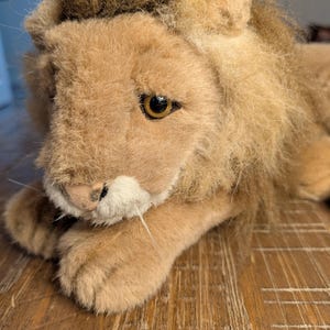 May include: A plush lion toy with a tan body and a brown mane. The lion has a detailed face with a white muzzle, whiskers, and dark eyes. The toy is lying down on a wooden surface.