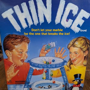 May include: A blue box for the "Thin Ice" game. The box features illustrations of children, marbles, and a penguin. Text on the box reads "Don't let your marble be the one that breaks the ice!" and "For 2 or more players Ages 5 and up."