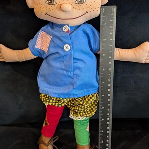 Op de afbeelding: Handgemaakte pop met een stoffen gezicht, sproeten en oranje haarvlechten. De pop draagt een blauw shirt, patchwork shorts en mismatched rode en groene sokken. De pop is ongeveer 30 cm groot.
