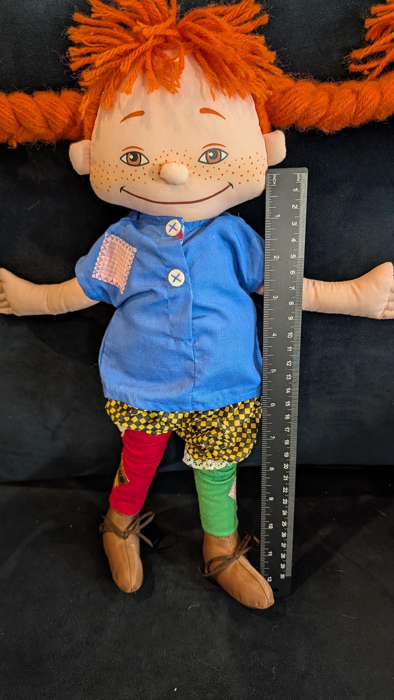 Op de afbeelding: Handgemaakte pop met een stoffen gezicht, sproeten en oranje haarvlechten. De pop draagt een blauw shirt, patchwork shorts en mismatched rode en groene sokken. De pop is ongeveer 30 cm groot.
