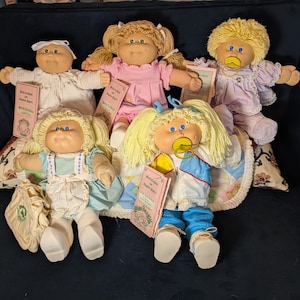 May include: Five Cabbage Patch Kids dolls are displayed, each with unique hairstyles and outfits. The dolls have soft, round faces, blue eyes, and some have pacifiers. Each doll holds a small pink book. The dolls are on a patterned surface.