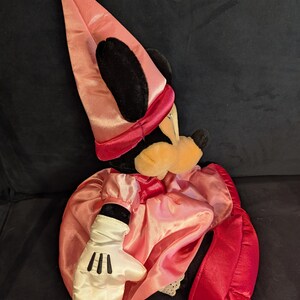 Peut inclure: Poup&eacute;e en peluche de Minnie Mouse habill&eacute;e en sorci&egrave;re. La poup&eacute;e porte une robe et un chapeau en satin rose, une cape en satin rouge et des gants blancs avec des d&eacute;tails noirs. Le visage de la poup&eacute;e a un nez jaune et des oreilles noires.