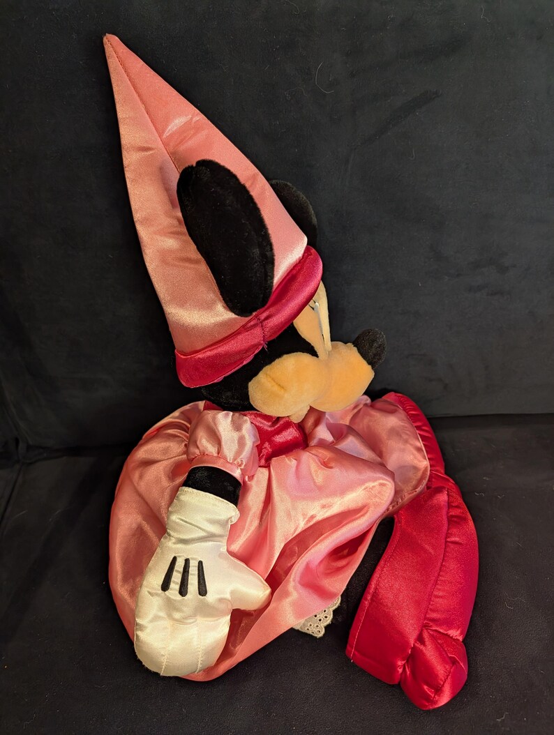 Peut inclure: Poup&eacute;e en peluche de Minnie Mouse habill&eacute;e en sorci&egrave;re. La poup&eacute;e porte une robe et un chapeau en satin rose, une cape en satin rouge et des gants blancs avec des d&eacute;tails noirs. Le visage de la poup&eacute;e a un nez jaune et des oreilles noires.