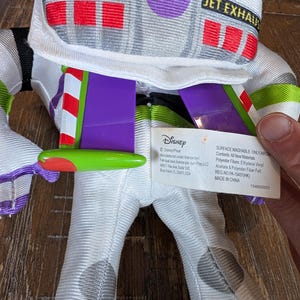 May include: A Buzz Lightyear plush toy, featuring a white and green spacesuit design. The toy has purple accents, red and white wings, and the text "DANGER!" and "JET EXHAUST" on the helmet. A Disney tag is visible.